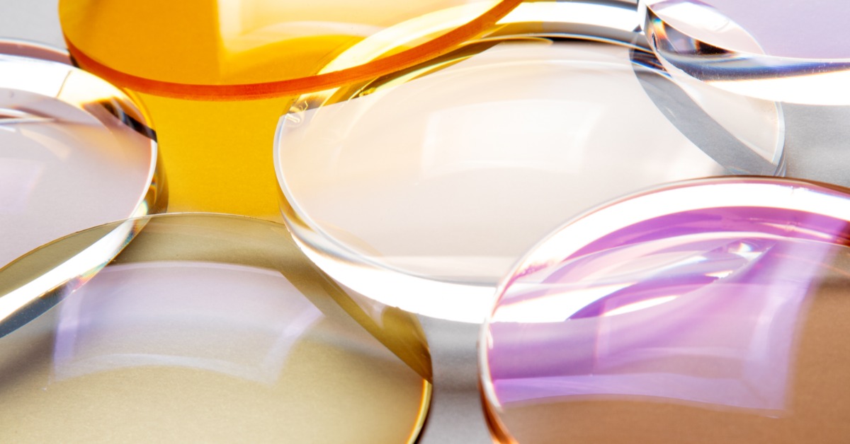 Close-up of overlapping optical glass lenses in various shapes and sizes, featuring transparent surfaces with yellow, purple, and amber tints.