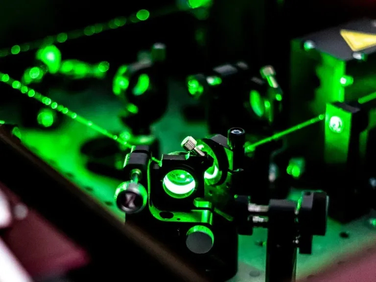A green laser beam crosses mirrors and mounts on an optical table inside a dark photonics laboratory.