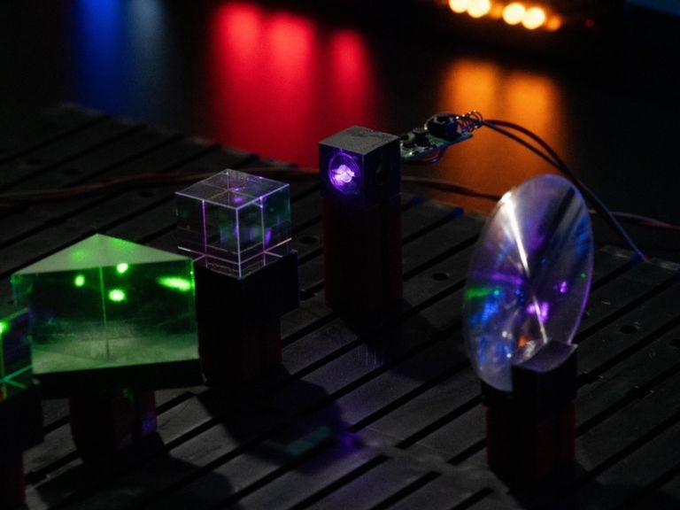 A lab setup contains green and purple lasers shinning through clear optical cubes and a rounded lens on a black table.