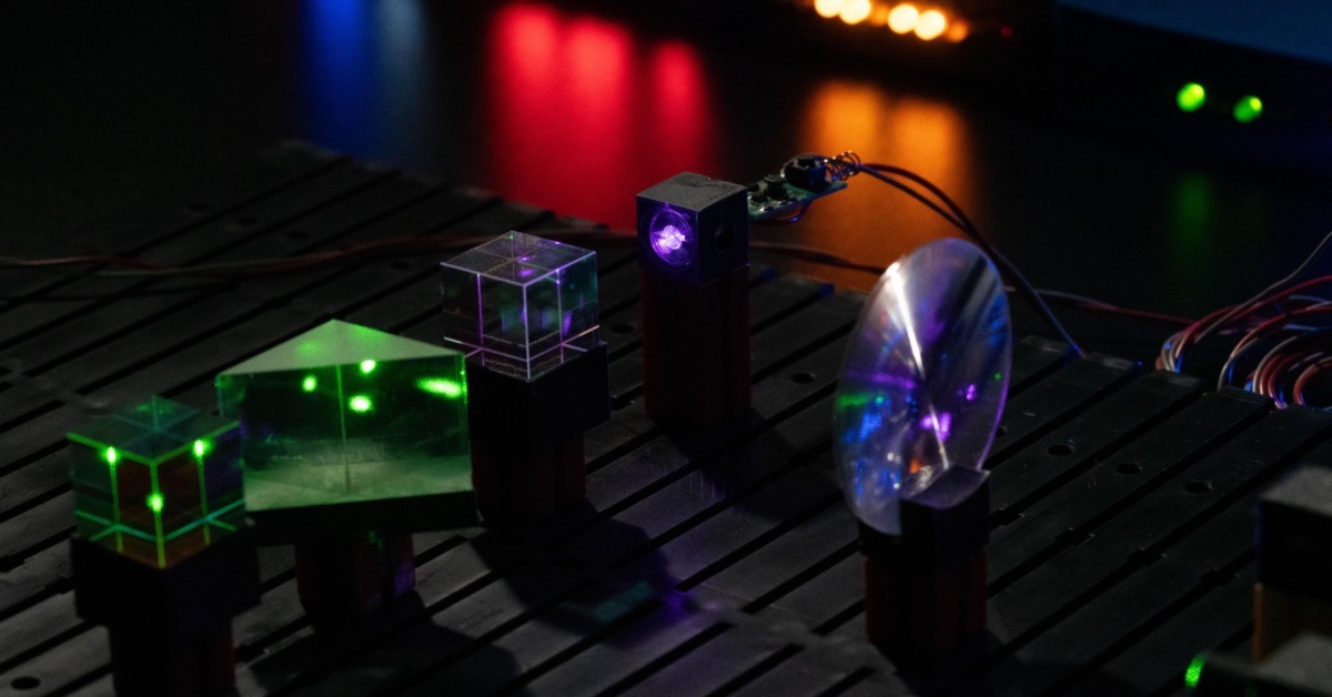 A lab setup contains green and purple lasers shinning through clear optical cubes and a rounded lens on a black table.